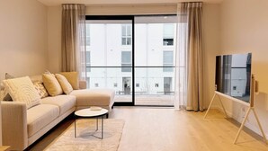 Ett modernt vardagsrum med en soffa, ett soffbord, en tv på ett tv-bord och stora fönster med gardiner.