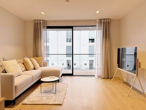 A modern living room with a sofa, a coffee table, a TV on a stand, and large windows with curtains. - Central Christchurch Apartment-Free Parking (Christchurch)