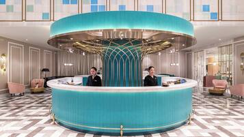 A hotel reception area with a central blue counter, two receptionists, and a check-in desk.