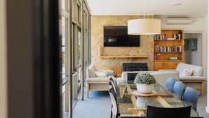 Een moderne woonkamer met een glazen eettafel, banken, een open haard, een televisie en een boekenkast.
