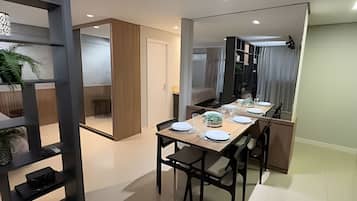 Un comedor moderno con una larga mesa dispuesta para cuatro personas, un espejo que refleja la estancia, y una planta en un estante.