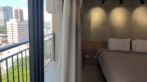 A hotel room with a bed, a nightstand, a window with curtains, and a view of city buildings. - Casa Confortavel em Foz do Iguaçu (Foz do Iguaçu)
