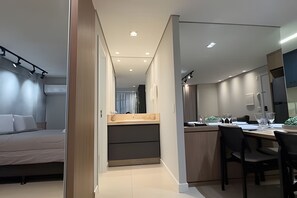 Una habitación de hotel moderna con cama, una pequeña kitchenette, un comedor con mesa y sillas, y un espejo grande.