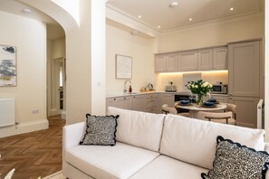 A modern kitchen with a dining area, a sofa with patterned cushions, and a hallway leading to another room. - Stunning 2 Bedroom Flat in the Centre of Cheltenham (Cheltenham)