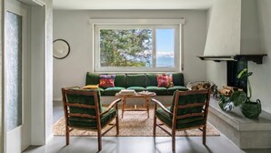 Ein Wohnzimmer mit einem grünen Sofa, zwei Holzsesseln, einem Couchtisch, einem Kamin und einem großen Fenster mit Blick auf Bäume.