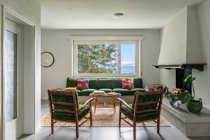 Ein Wohnzimmer mit einem grünen Sofa, zwei Holzsesseln, einem Couchtisch, einem Kamin und einem großen Fenster mit Blick auf Bäume.
