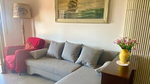 En stue med en grå sofa, en rød lænestol, en lampe, et billede af et skib i ramme og en vase med blomster.