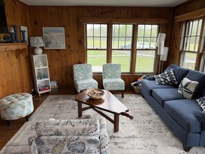 A cozy living room with wooden paneling, a blue sofa, floral-patterned chairs, a wooden coffee table, and a view of the outdoors through large windows.