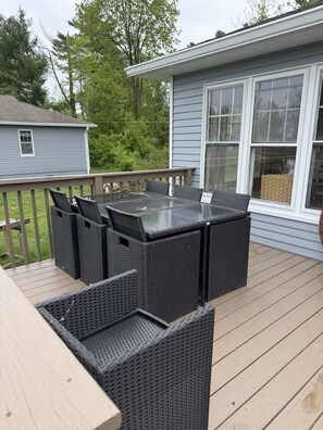 A deck with a glass-top table and wicker chairs, overlooking a house with large windows.
