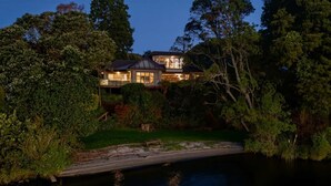 A large house with a deck, surrounded by dense trees and a body of water. - Lion House - Lakefront Spa, Private Beach & Jetty (Rotoiti Forest)
