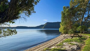 A lakeside view with a picnic table, trees, and a distant mountain range. - Lion House - Lakefront Spa, Private Beach & Jetty (Rotoiti Forest)