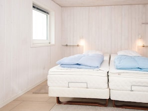 A room with two beds, each with a mattress and pillows, a window, and wall-mounted lighting.