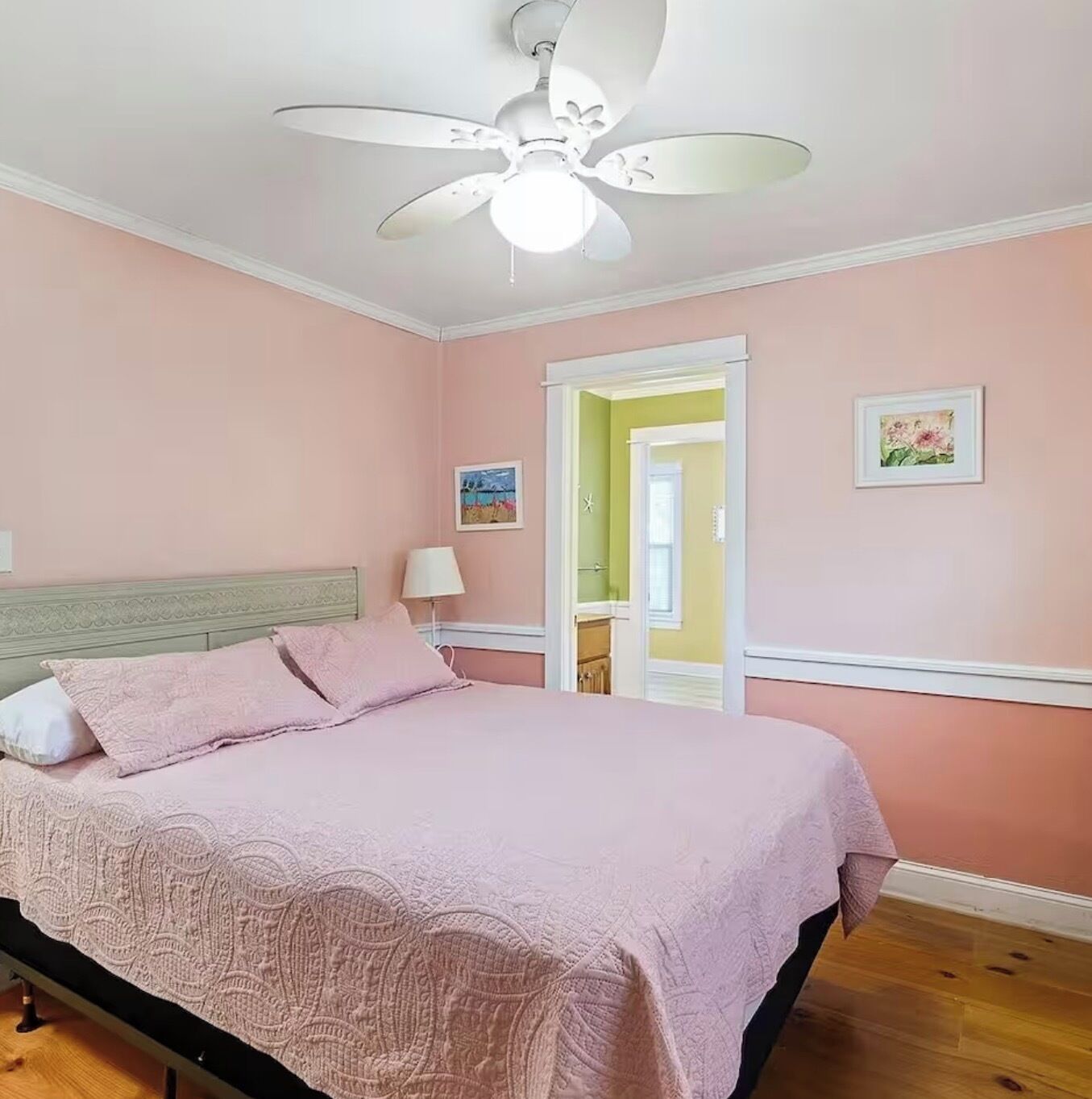 A bedroom with a bed, a ceiling fan, a door leading to another room, and a wall with a picture.