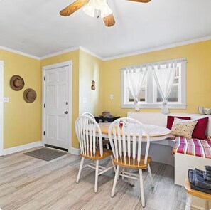 A room with a dining table, two white chairs, a sofa with a colorful blanket, a window with white curtains, and a door. - 3-bedroom cheerful cottage in pleasant Carolina Beach! (Carolina Beach)