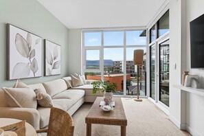 A living room with a sofa, a coffee table, and a large window with a view of buildings and mountains.