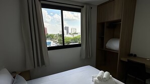 A hotel room with a bed, a window with curtains, a wooden wardrobe, and a view of buildings. - Casa Confortavel em Foz do Iguaçu (Foz do Iguaçu)