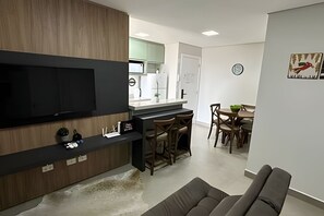 A modern living room with a flat-screen TV, wooden paneling, a dining area with a table and chairs, and a kitchen in the background. - Casa Confortavel em Foz do Iguaçu (Foz do Iguaçu)