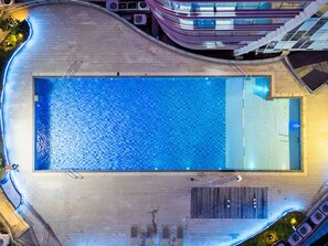 Ein rechteckiger Swimmingpool mit glasklarer, blauer Wasseroberfläche, umgeben von einer Betonplattform und angrenzenden Gebäuden.