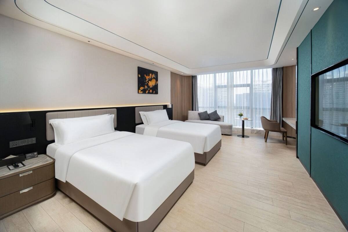 A modern hotel room with two beds, a desk, a chair, and a view of the city.