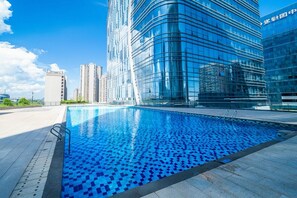 Um edifício moderno com fachada de vidro refletor, piscina com azulejos azuis em mosaico e céu claro.