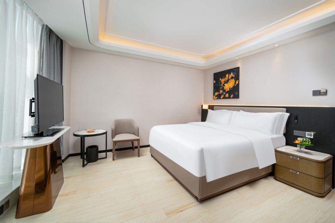 Quarto de hotel com cama, televisão, pequena mesa, cadeira e mesa de cabeceira com abajur.