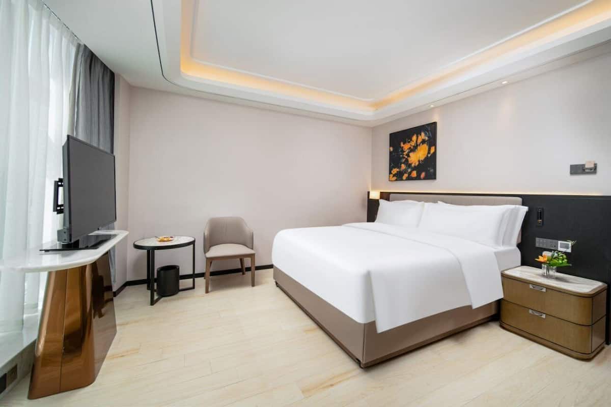 A hotel room with a bed, a TV, a small table, a chair, and a nightstand with a lamp.