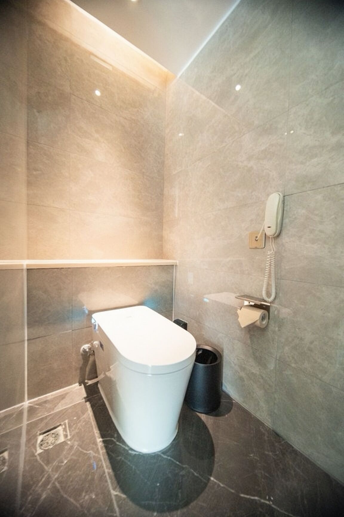 A modern bathroom with a toilet, a trash bin, and a telephone mounted on the wall.