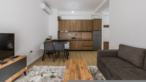 Un soggiorno moderno con un divano, un tavolino da caffè e una televisione. È presente un angolo cottura con mobili in legno e un tavolo da pranzo con sedie.