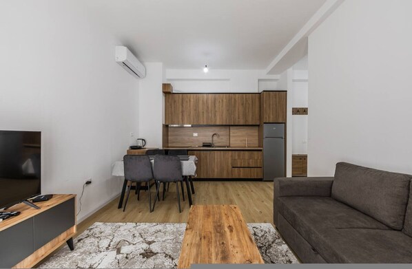 A modern living room with a sofa, a coffee table, and a TV. There is a kitchen area with wooden cabinets and a dining table with chairs. - Central Quiet Tirana Flat 1BR Apartment (Tirana)
