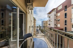 A balcony with a chair, a glass door, and a view of a street with parked cars and buildings. - Sunset Apartment 1BR Escape (Durrës)