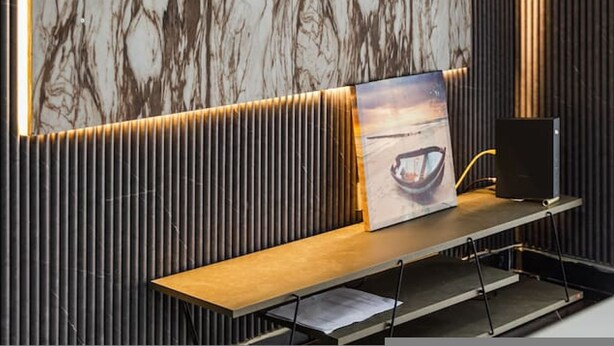 A modern interior with a flat-screen TV mounted on a marble-patterned wall, a wooden bench with a picture frame, and a speaker on a stand. - Sunset Apartment 1BR Escape (Durrës)