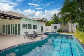 A swimming pool with tiled edges, a covered patio with lounge chairs, and a house with large windows. - Orchid Paradise Homes OPV307 (Tambon Hin Lek Fai)