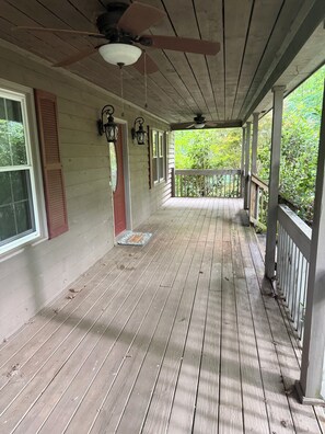 A wooden porch with a ceiling fan, a welcome mat, and a view of greenery.