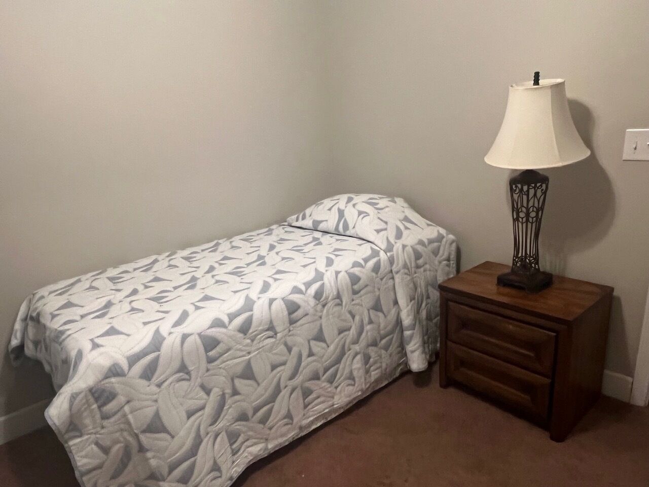 Ein Einzelbett mit einem gemusterten Bettüberwurf, ein Nachttisch mit Lampe und eine schlichte Wand.