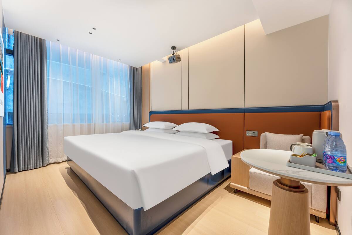 Una habitación de hotel moderna con una cama grande, una mesita pequeña, un sofá y un ventanal con cortinas.
