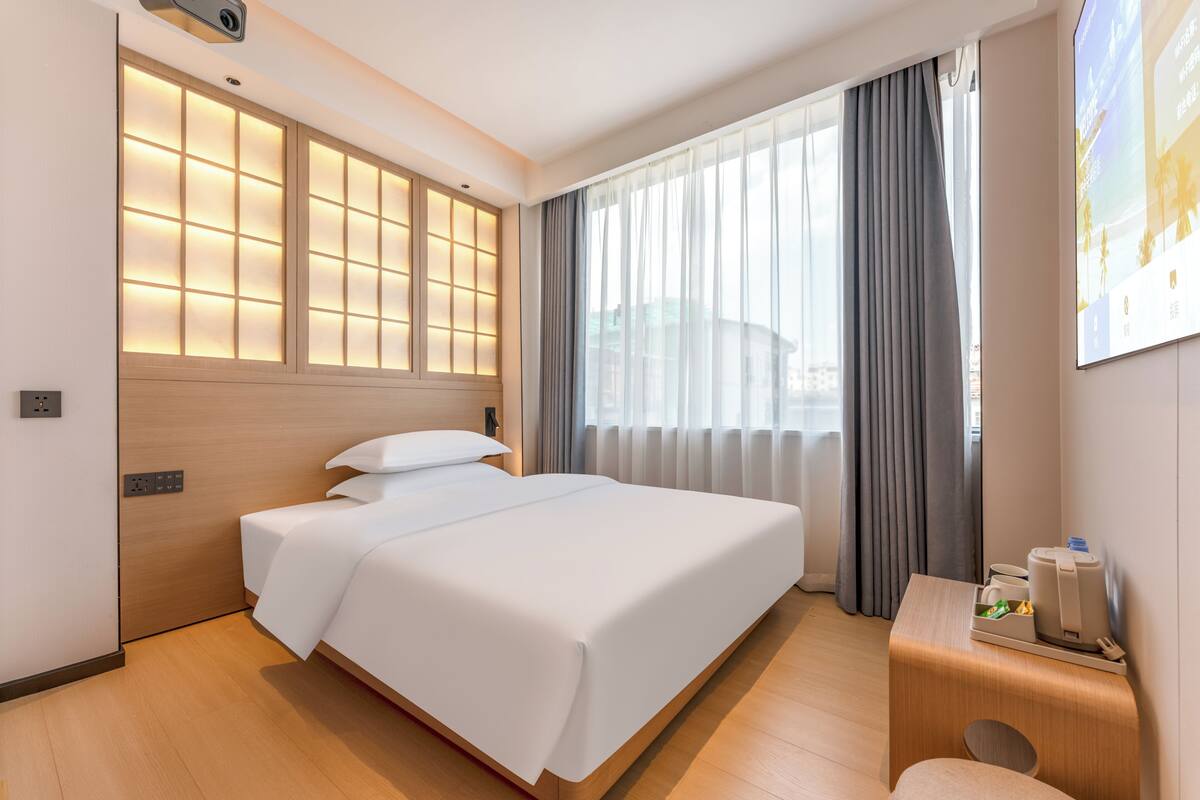 Una habitación de hotel con cama, escritorio con lámpara, ventana con cortinas y televisor instalado en la pared.