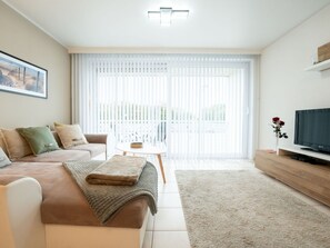 Ein modernes Wohnzimmer mit einer Couch, einem Couchtisch, einem Fernseher und einem großen Fenster mit Jalousien.