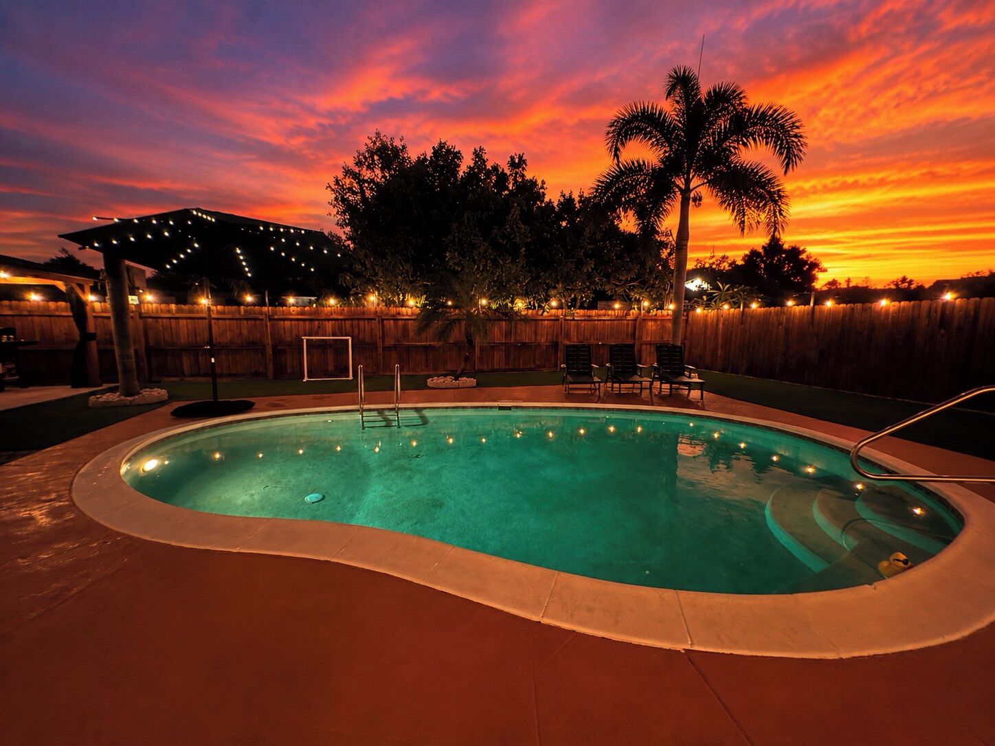 Une piscine avec une terrasse, des palmiers et un coucher de soleil.