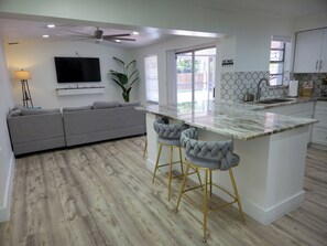 A modern kitchen with a central island, bar stools, a grey sofa, and a ceiling fan. - Velara Naples, Beautiful Family Pool Home Near Beach & Downtown. (Naples)