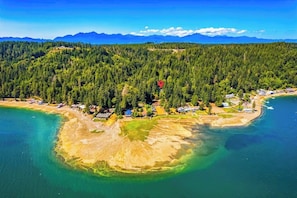 An aerial view of a coastal area with a forested hill, a beach, and a small settlement with houses and boats.