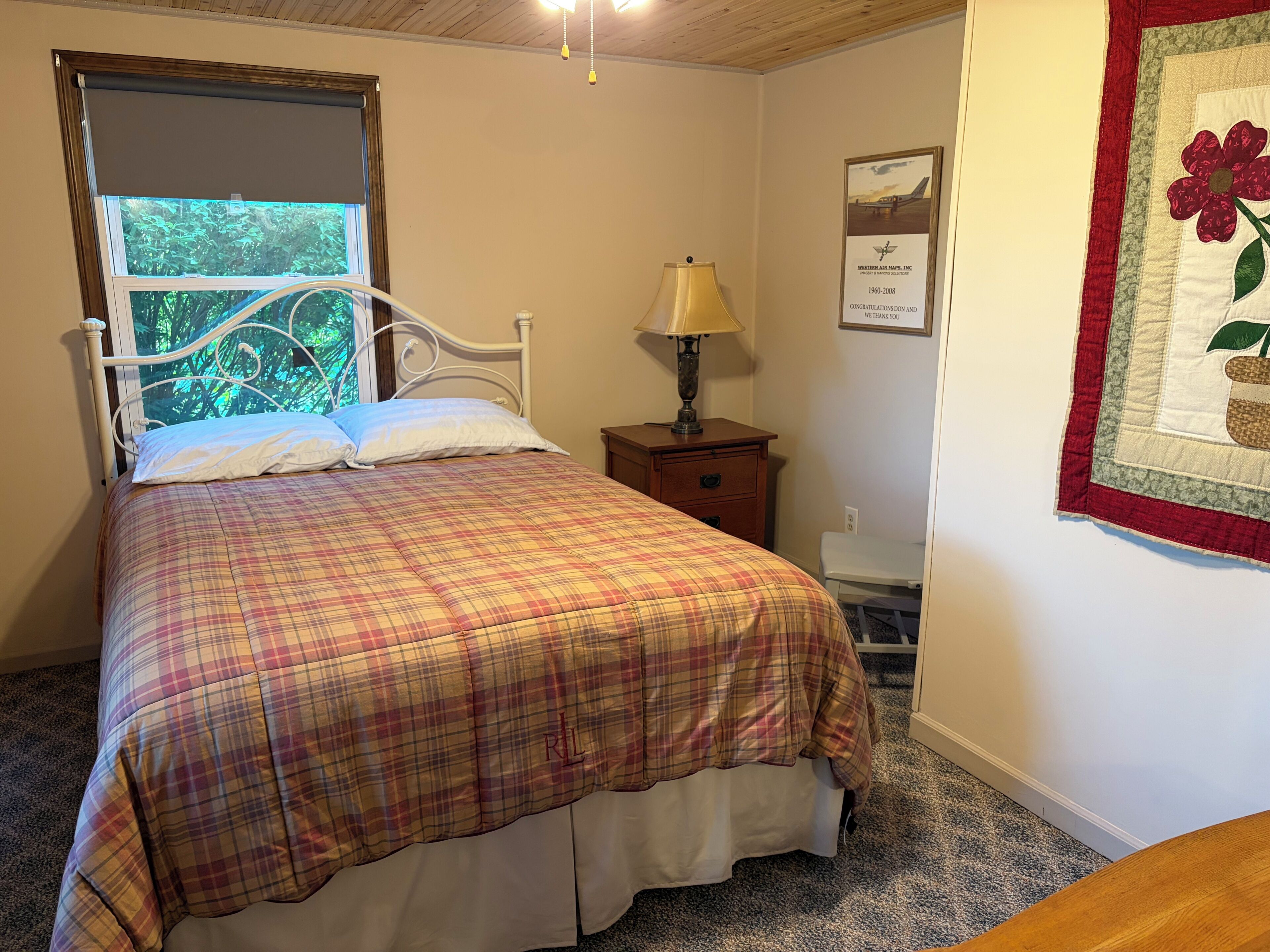 A bedroom with a bed, a nightstand, a lamp, a window with a view, and a framed certificate on the wall.