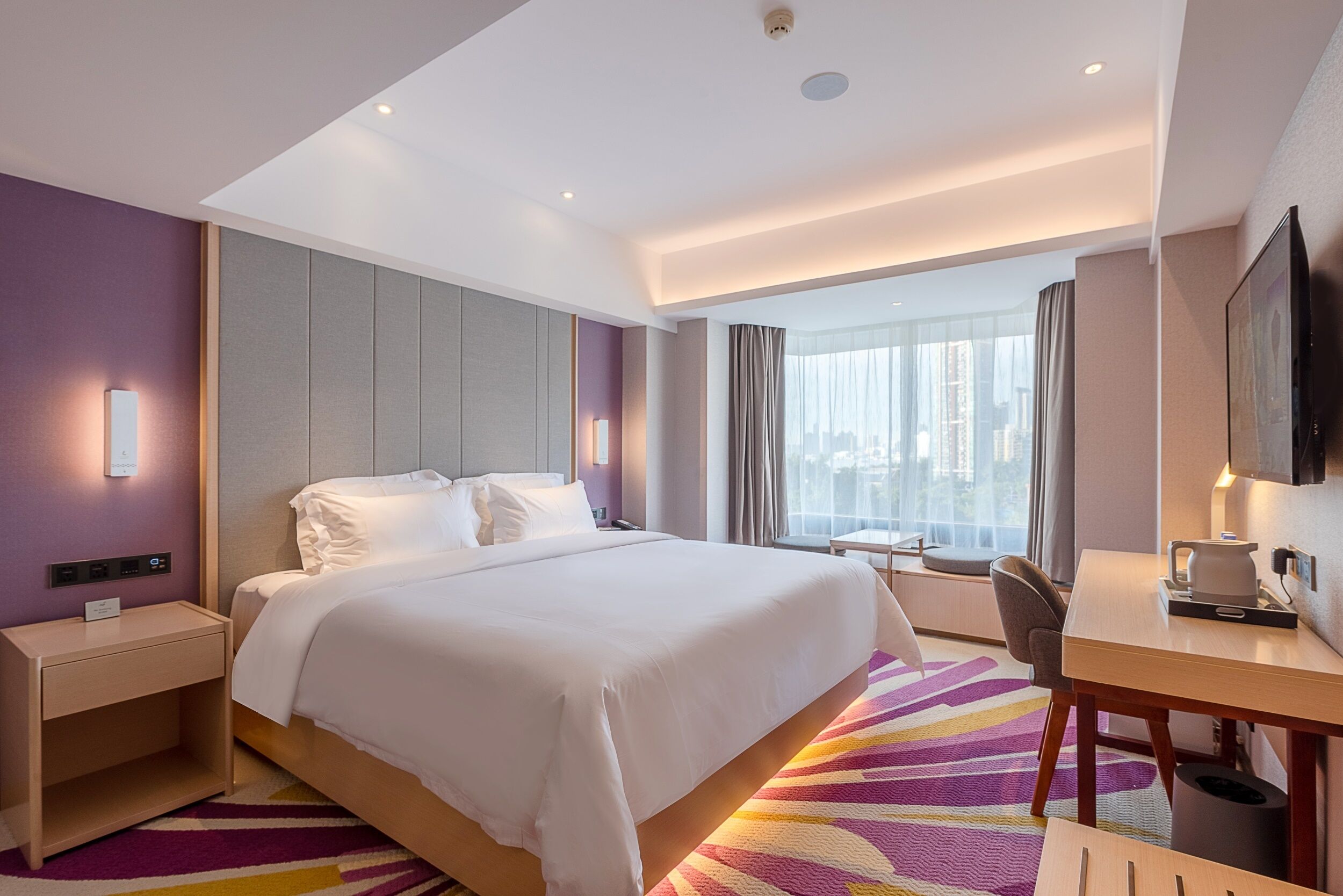 Une chambre d’hôtel équipée d’un lit, d’une table de chevet, d’un bureau, d’une chaise et d’une grande fenêtre donnant sur la ville.