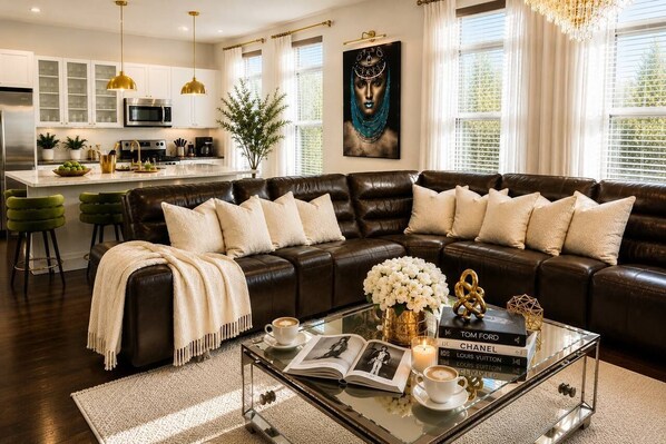 A modern living room with a leather sofa, a glass coffee table with books and a candle, and a kitchen in the background.