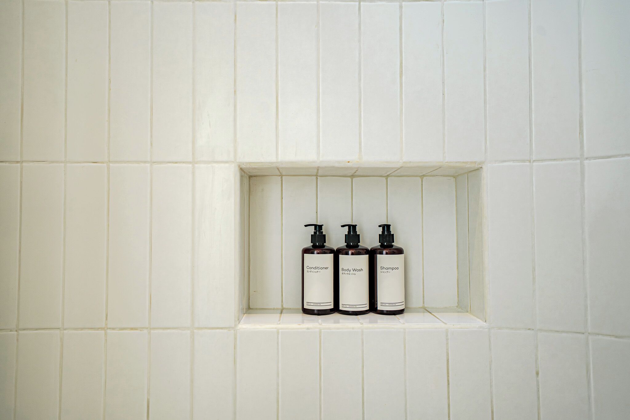 Un baño con paredes de azulejos blancos y un estante con tres artículos de aseo.