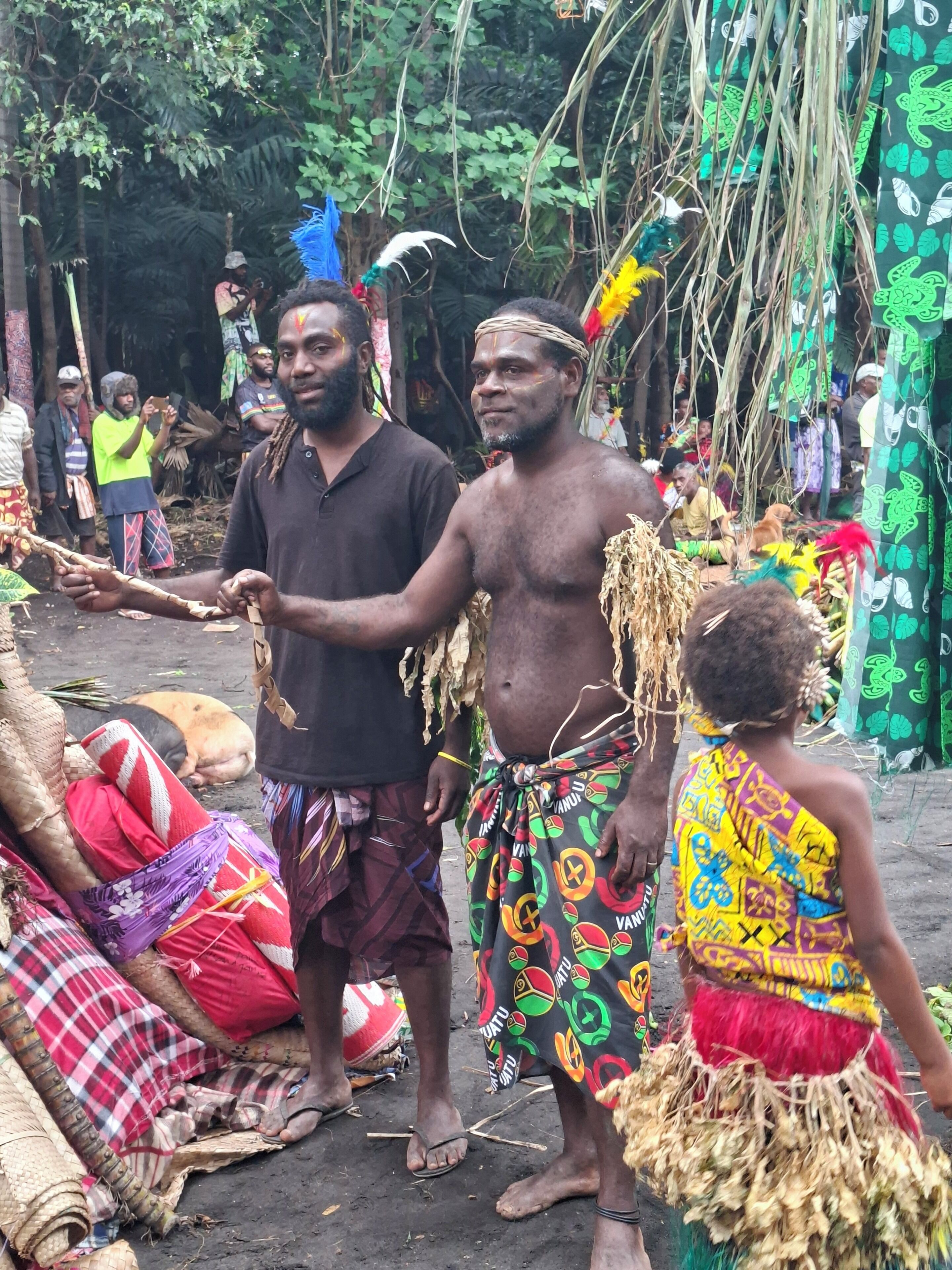 Un grupo de personas con vestimenta tradicional, algunas adornadas con plumas y cuentas, reunidas en un entorno al aire libre con árboles y un refugio improvisado al fondo.