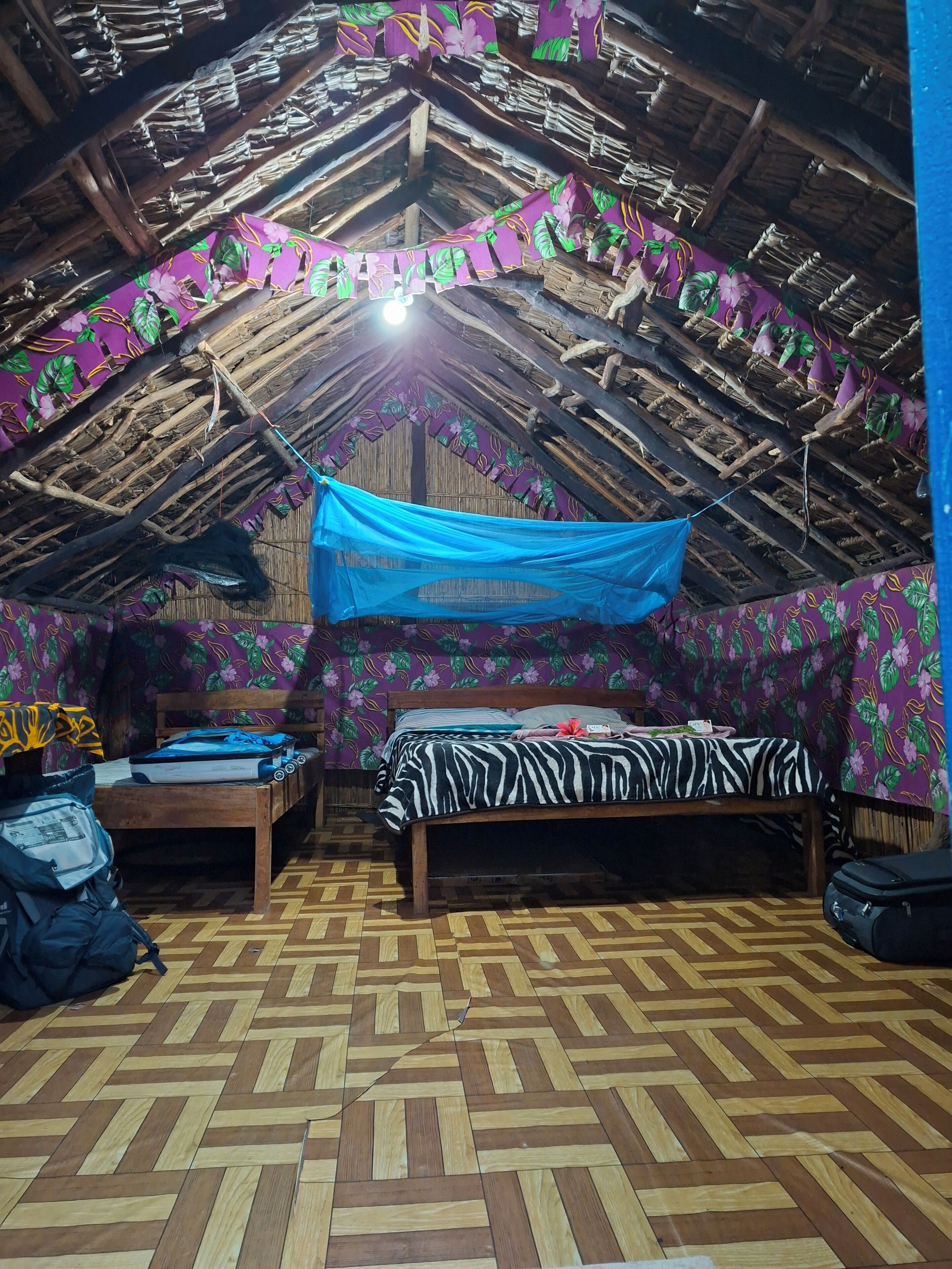 Um quarto com telhado de palha, cama com colcha de estampa de zebra, piso de madeira e um tecido azul pendurado.