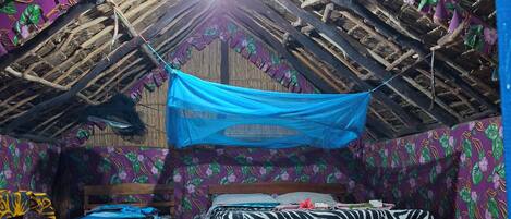 Um quarto com telhado de palha, cama com colcha de estampa de zebra, piso de madeira e um tecido azul pendurado.