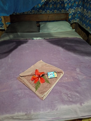 A bed with a purple bedspread, a folded towel with a hibiscus flower, and a box of Giv brand deodorant.