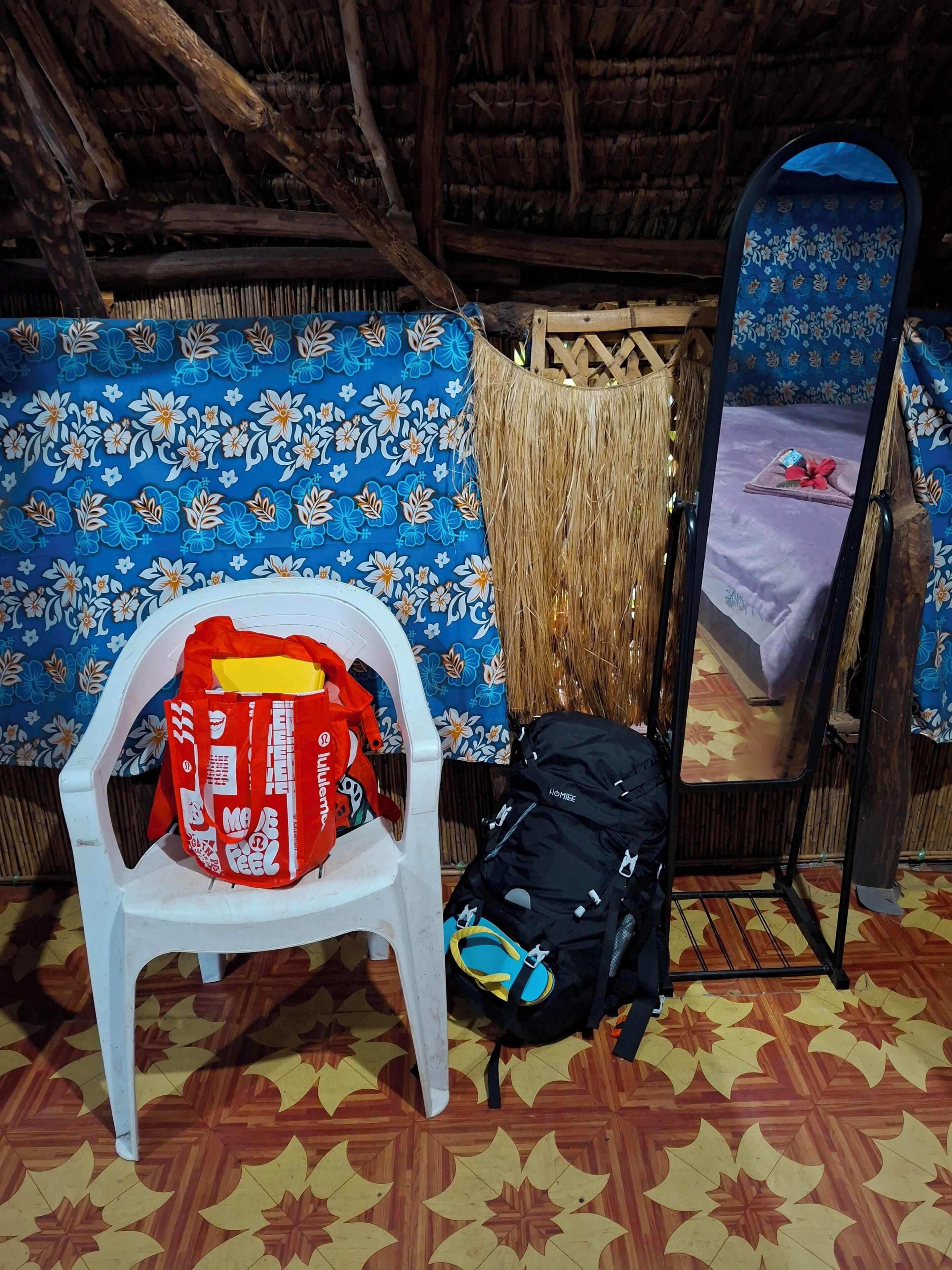 Una habitación con techo de paja, una cama con colcha de motivos florales, una silla de plástico blanco y una mochila negra en el suelo.