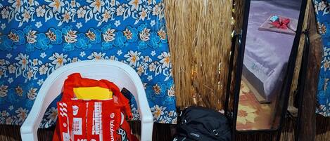Una habitación con techo de paja, una cama con colcha de motivos florales, una silla de plástico blanco y una mochila negra en el suelo.